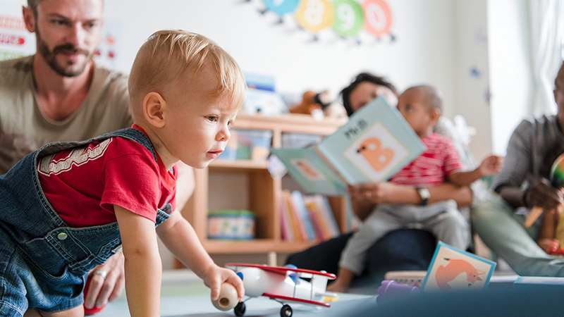 Kleiner Bub spielt im Kindergarten am Boden. Pädagoginnen sind vor Ort.