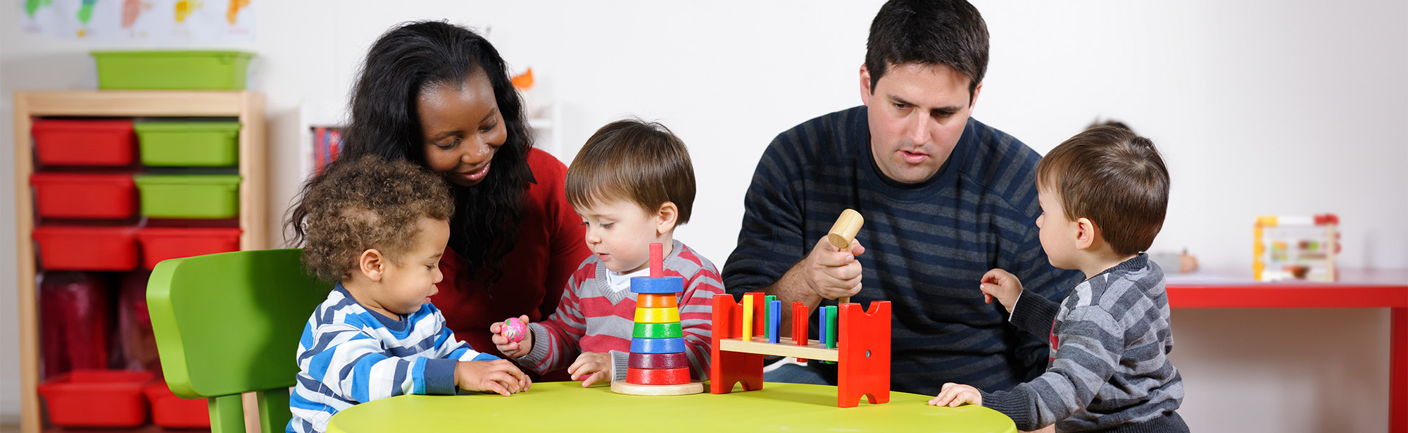 Weibliche Pädagoging und männlicher Pädagoge mit 3 Kindern und Spielzeug an einem Tisch